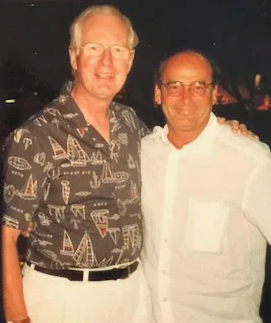 Picture of Merrick and Don smiling for the camera. Merrick wears a patterned short-sleeved shirt and Don a white one. They both have their arms over each other's shoulders.